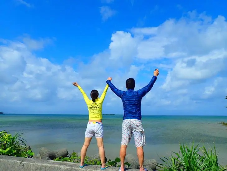 青い海に向かって両手を広げ、ラッシュガードを着て立つ男女の後ろ姿。