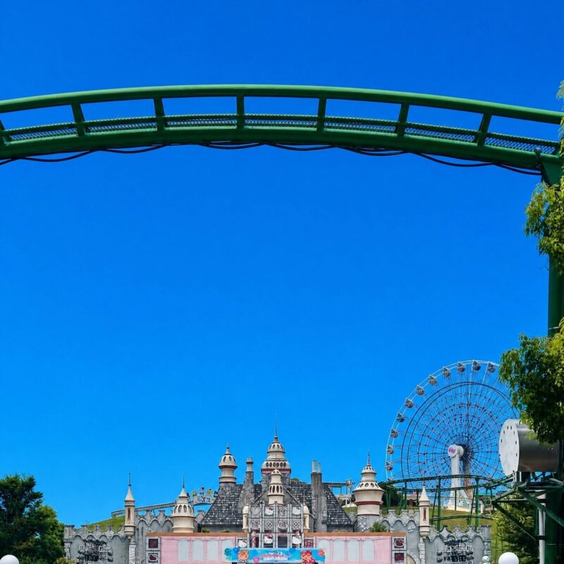 快晴の青空の下、ジェットコースターの緑色のレールアーチ越しにハーモニーランドのお城と観覧車が見える風景です。