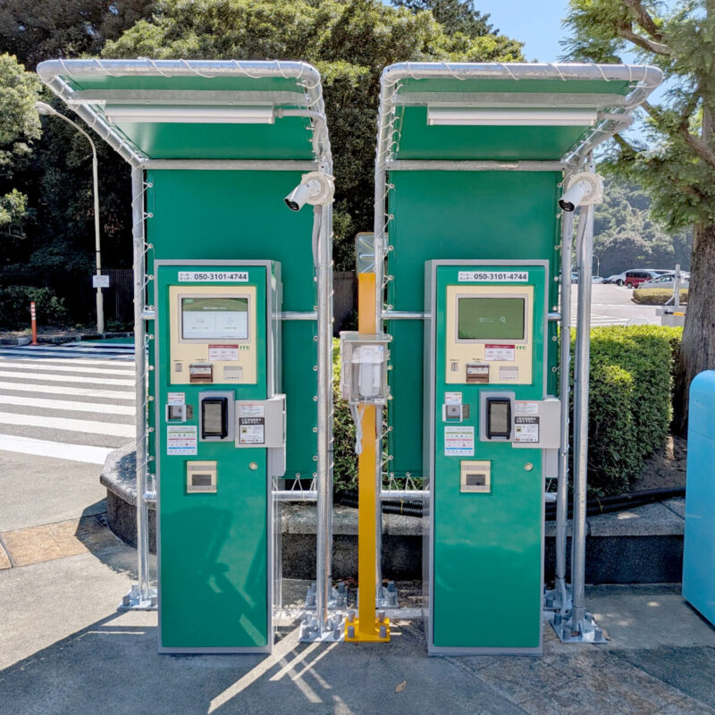 屋外に設置された緑色の自動精算機が2台並んでいます。上部には雨除けの屋根と防犯カメラが取り付けられています。